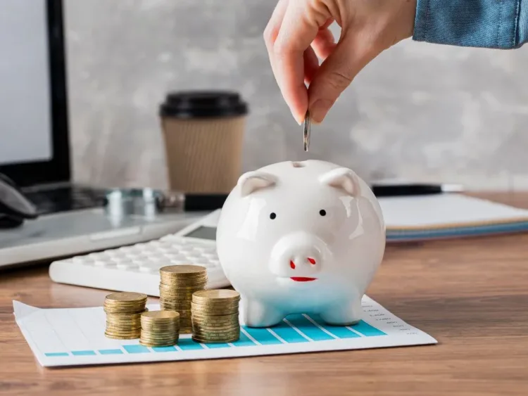 Mão colocando dinheiro no cofrinho, com calendário e calculadora completando a imagem.